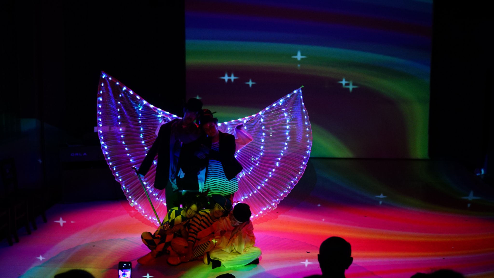 Zdjęcie prezentuje grupę ludzi znajdujących się na scenie w kolorowym świetle scenicznym