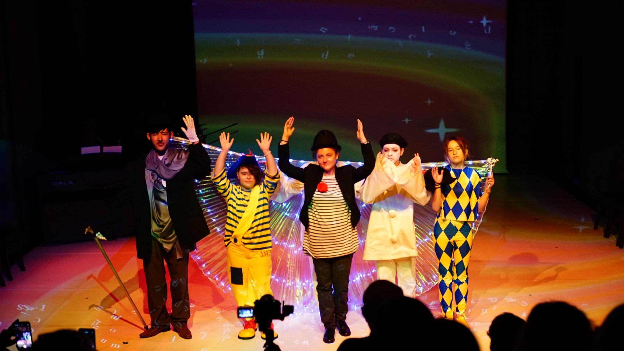Zdjęcie prezentuje grupę ludzi znajdujących się na scenie w kolorowym świetle scenicznym