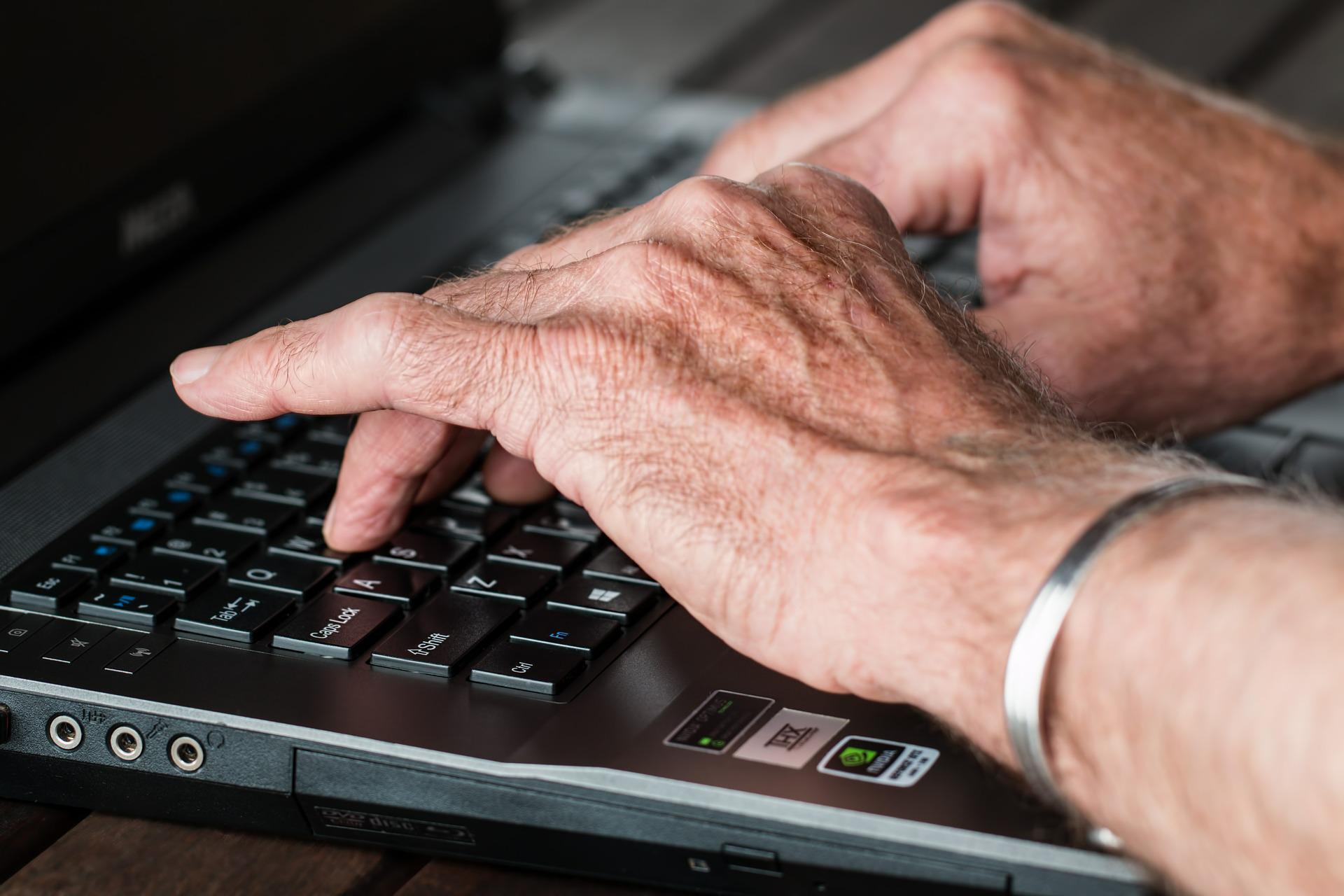 fotografia przedstawia ręcę starszej osoby piszącej coś na klawiaturze laptopa. Dłonie są pomarszczone, a na lewym nadgarstku znajdue się metalowa bransoletka. Laptop jest koloru czarnego i widać jedynie fragment klawiatury i skrawek monitora.