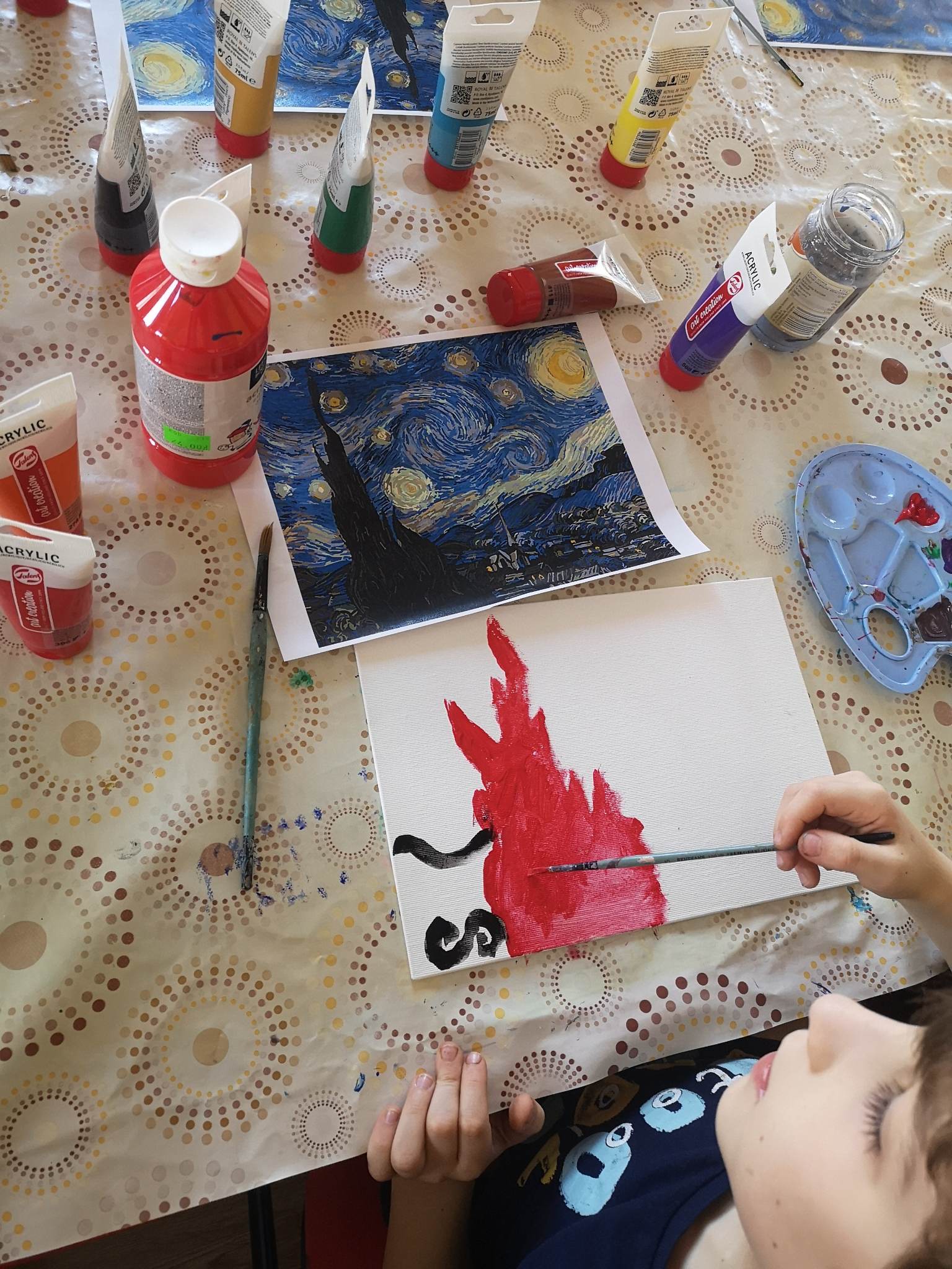 Zdjęcie przedstawia pracę malarską, która odmalowywana ze wzoru obrazu van Goga jest malowana przez nieujętą w kadrze zdjęcia osobę. Obraz van Goga to czarna wieża na tle niebieskim z żółtymi okręgami na niebie, a praca odmalowywana to czerwony ogień w kształcie wieży van Goga z dwoma czarnymi pasami po lewej stronie płótna