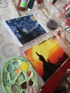 Zdjęcie przedstawia pracę malarską, która odmalowywana ze wzoru obrazu van Goga jest malowana przez nieujętą w kadrze zdjęcia osobę. Obraz van Goga to czarna wieża na tle niebieskim z żółtymi okręgami na niebie, praca odmalowana jest innej kolorystyki na tle pomarańczowym, żółtym i czerwonym.