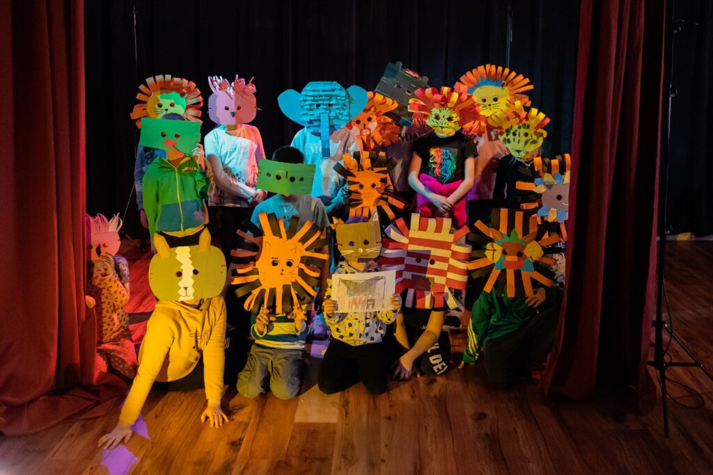 zdjęcie przedstawiające dzieci podświetlone reflektorem scenicznym na scenie, ubrane w maski, zakrywające ich twarze. Maski przedstawiają różne zwierzęta takie jak słonie, lwy, niedźwiedzie