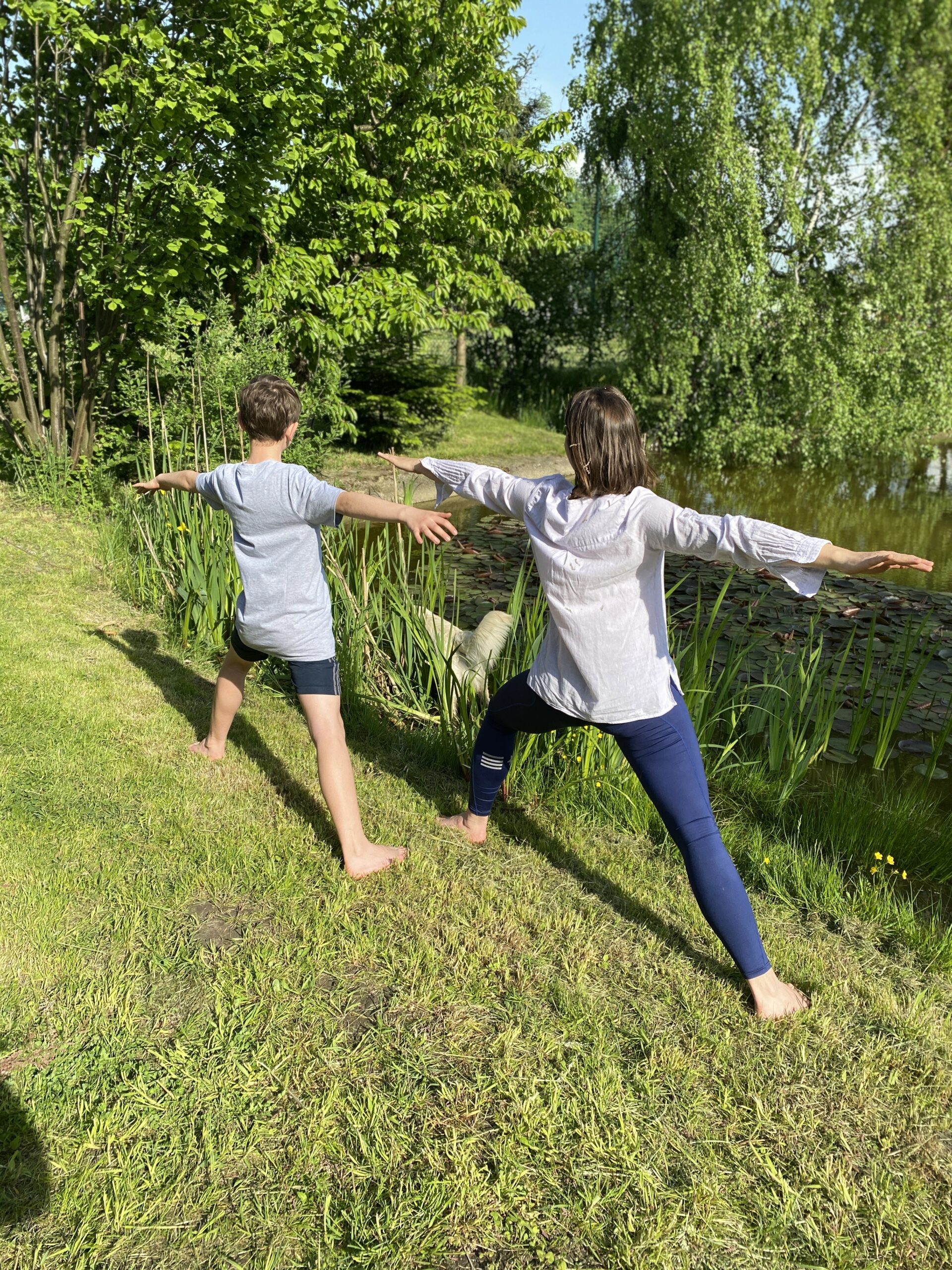 Fotografia przedstawia kobietę i chłopca stojących w postawie z jogi z rozchylonymi na boki rękami i ugiętym lewym kolanem, a prawa noga jest wyprostowana. Wykonują tę pozę na łonie natury tuż obok małego stawu stojąc na boso na trawie