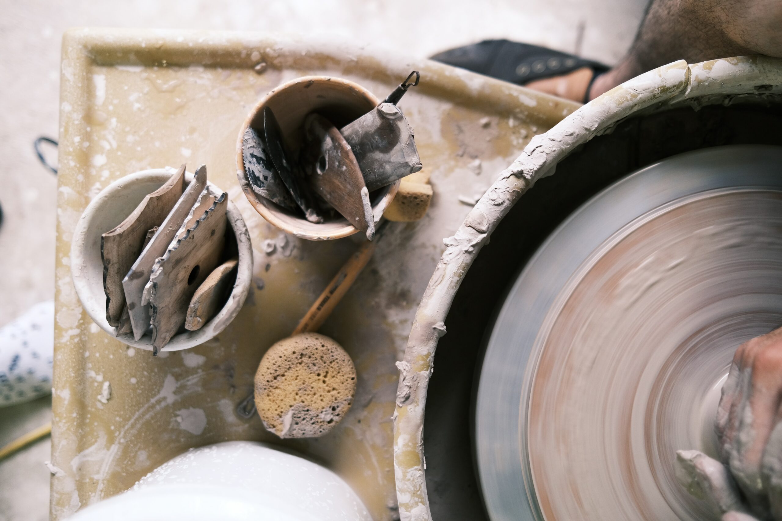 Zdjęcie przedstawia małe prace z ceramiki stojące na blacie obok koła garncarskiego w trakcie pracy. Prace ceramiczne są nieskończone