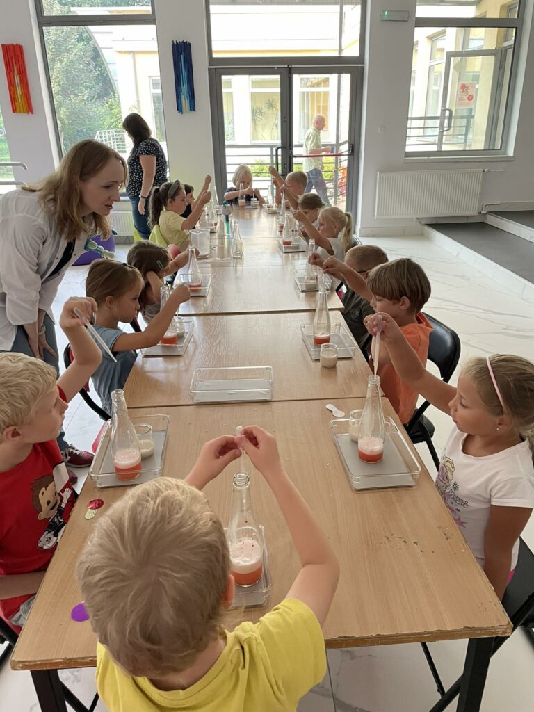 grupka dzieci wraz z prowadzącą przeprowadzają eksperymenty chemiczne przy długim stole